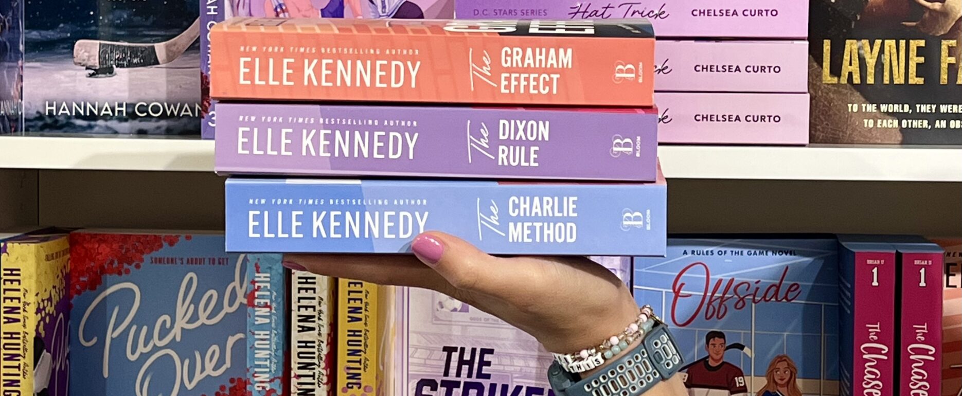 Holding a stack of 3 books by Elle Kennedy in front of a bookshelf