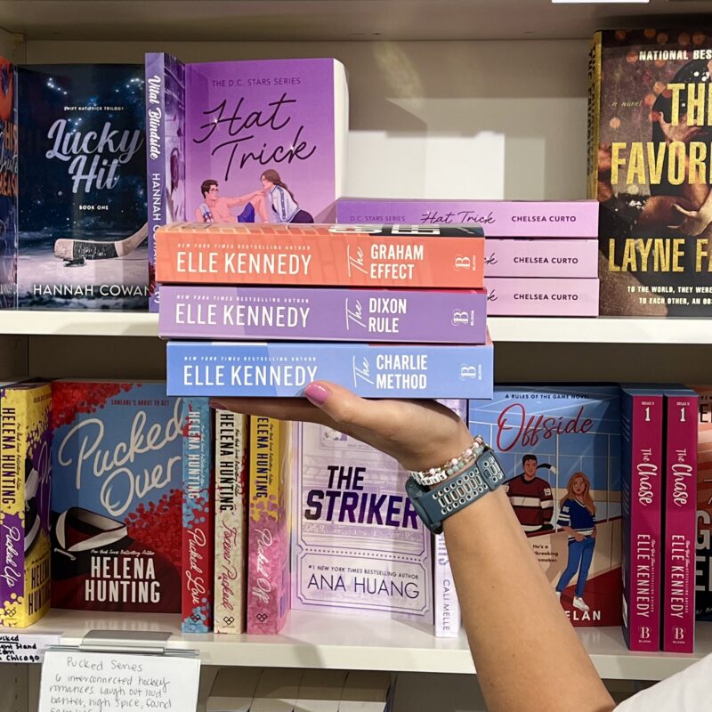 Holding a stack of 3 books by Elle Kennedy in front of a bookshelf