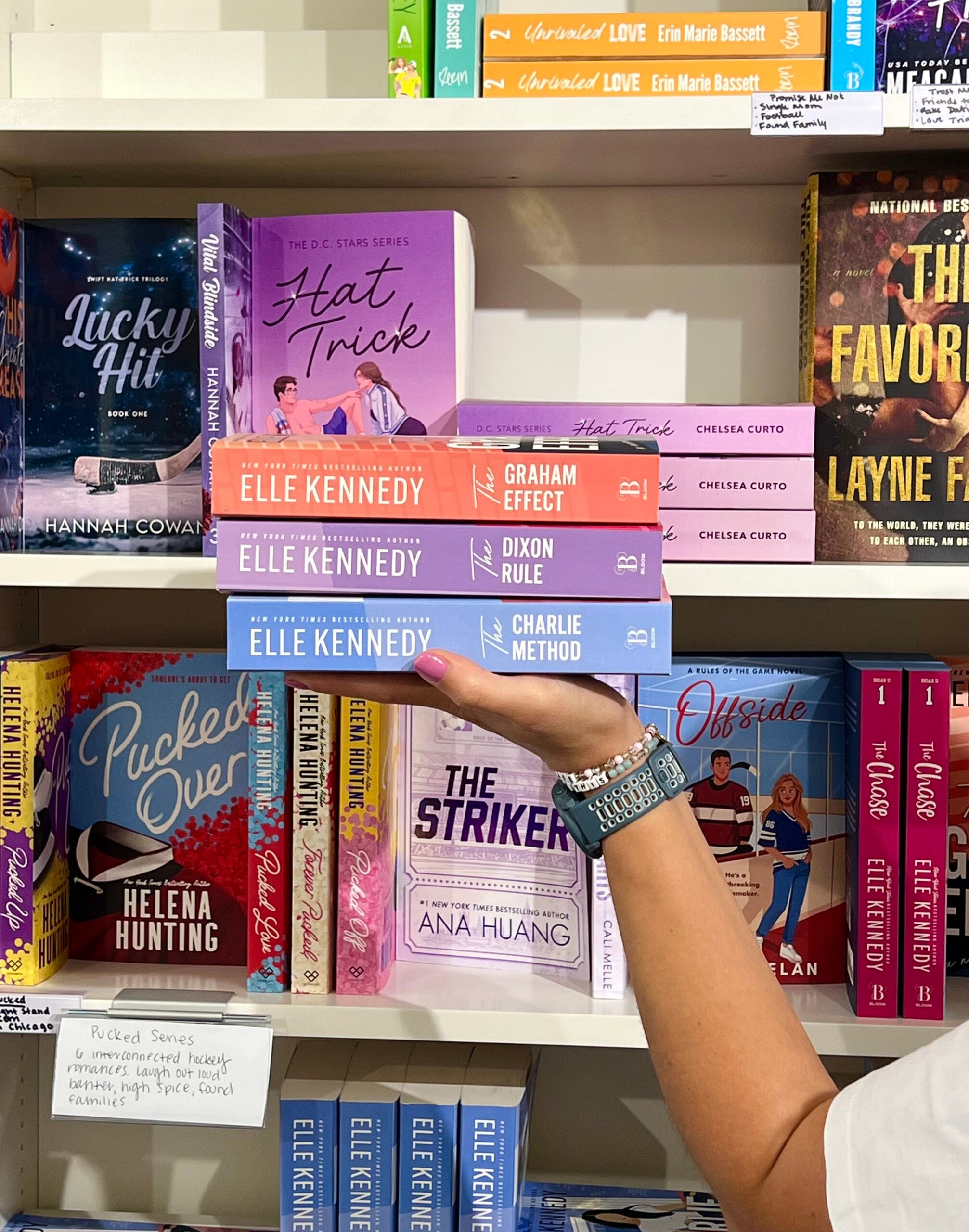 Holding a stack of 3 books by Elle Kennedy in front of a bookshelf
