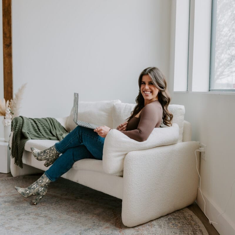Woman with brown hair sitting on a white couch with her legs crossed, smiling at the camera.
