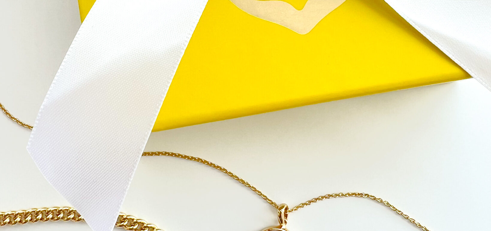 uplclose image of a kendra scott yellow jewlwelry box with a white ribbon and 3 gold necklaces, one with a green charm, gold chain, and an A charm