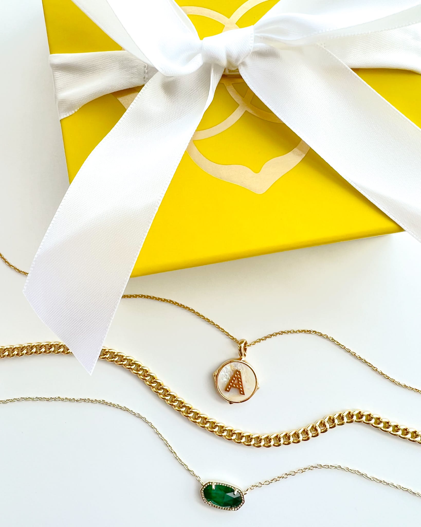 uplclose image of a kendra scott yellow jewlwelry box with a white ribbon and 3 gold necklaces, one with a green charm, gold chain, and an A charm