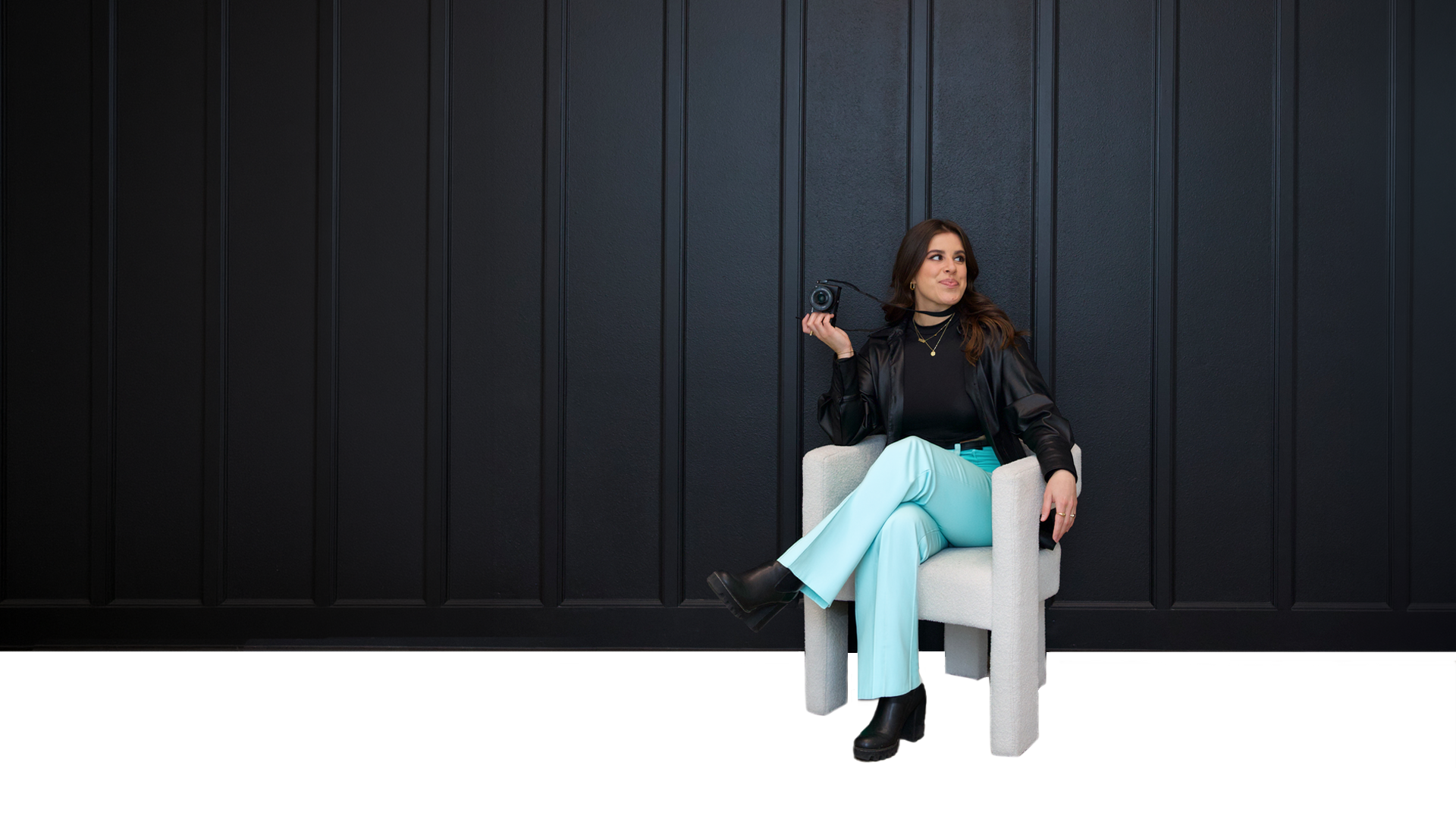 Social Media Manager woman with brown hair, black jacket, blue pants, sitting in a white chair smiling holding up a camera